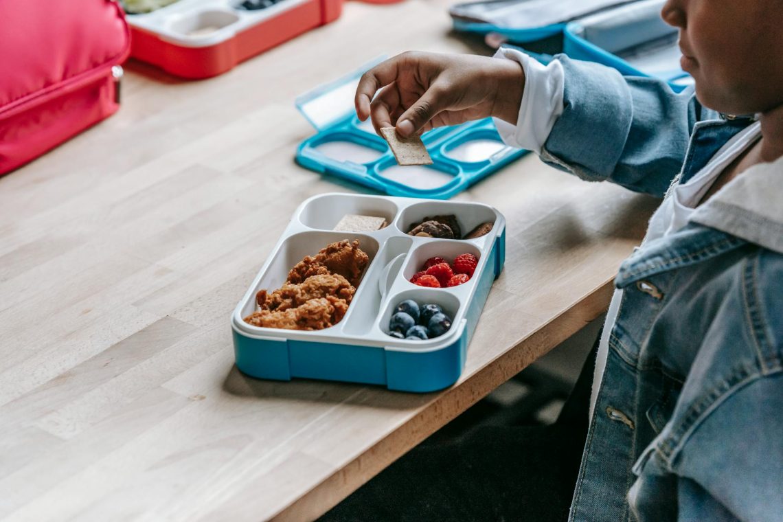 crop black schoolchild with biscuit near lunch box